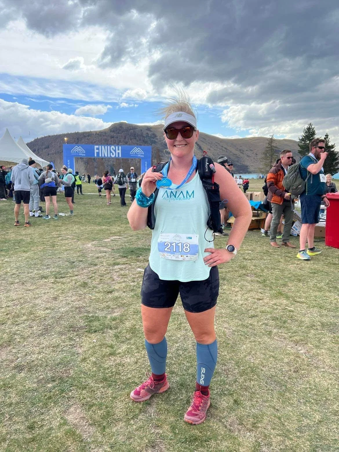 A smiling woman in running gear holding a finisher medal at a race event with a 'Finish' arch and other runners in the background.