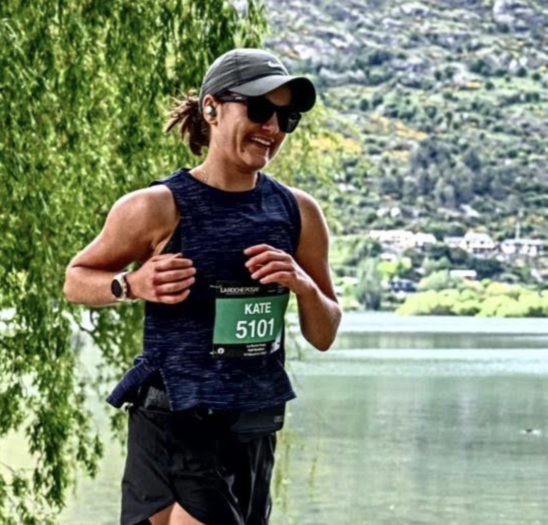 A woman running outdoors near a lake, wearing a dark athletic tank top, black shorts, sunglasses, a cap, and a race bib with the name "Kate" and number "5101."