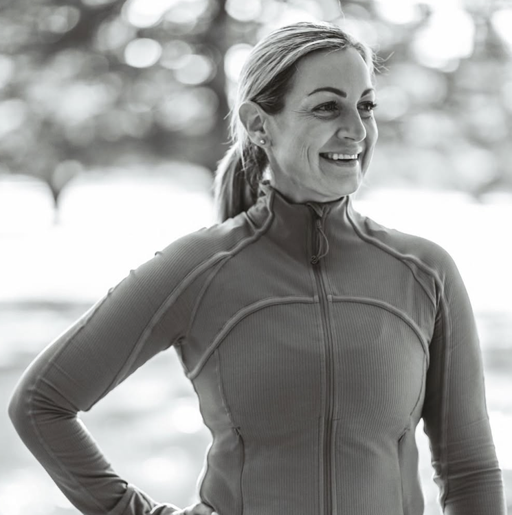A woman with blonde hair tied back, smiling, wearing a athletic zip-up jacket, outdoors with blurred trees in the background.