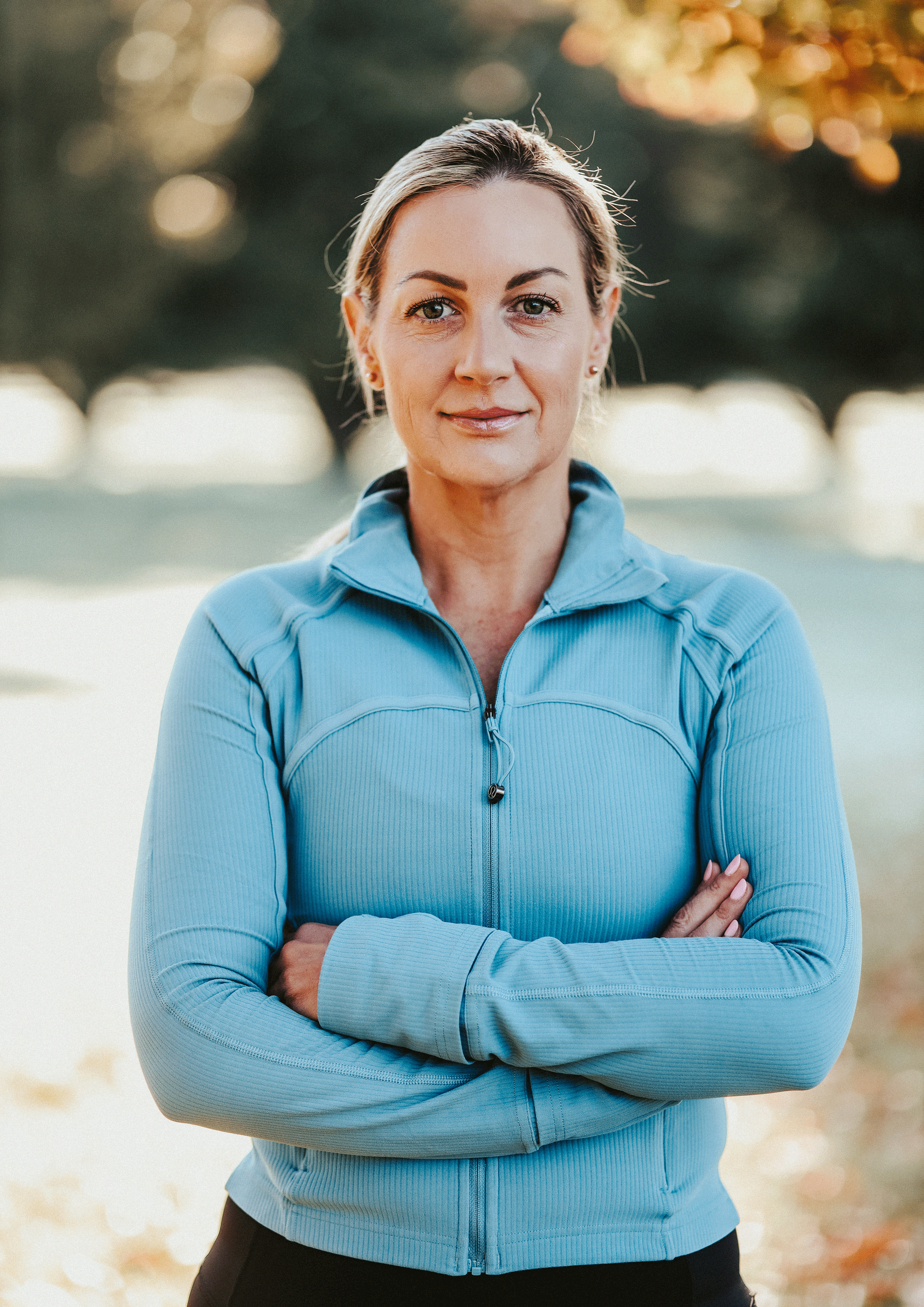 A woman in a light blue athletic jacket standing outdoors with her arms crossed, background of trees with fall foliage.