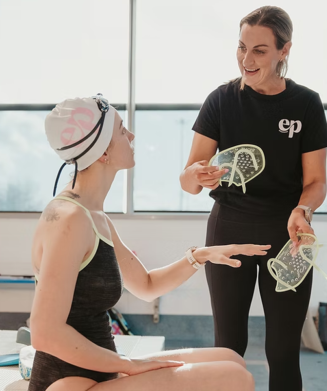 A woman healing a woman with a wearable medical device, seated and wearing a cap with electrodes and a strap, while the standing woman holds a device with wires in a medical or therapy setting with a bright window in the background.