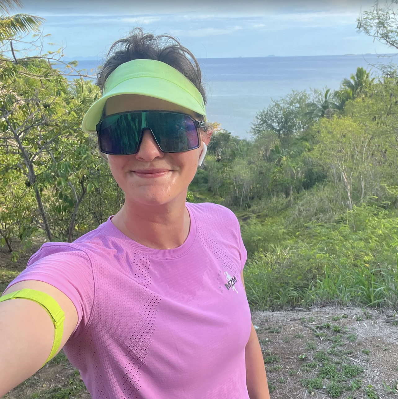 Woman taking a selfie outdoors, wearing a bright yellow visor, large black sunglasses, a pink athletic shirt, and a yellow wristband, with greenery, trees, and a body of water in the background.