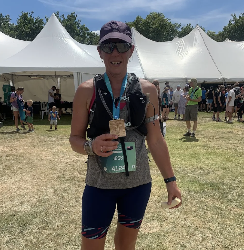 A woman at a race event holds a medal and a banana, standing outdoors in front of a large white tent surrounded by other people, some wearing race gear.
