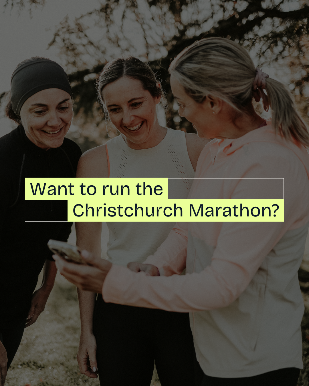 Three women smiling and looking at a phone outdoors during the daytime with trees in the background, promoting the Christchurch Marathon.