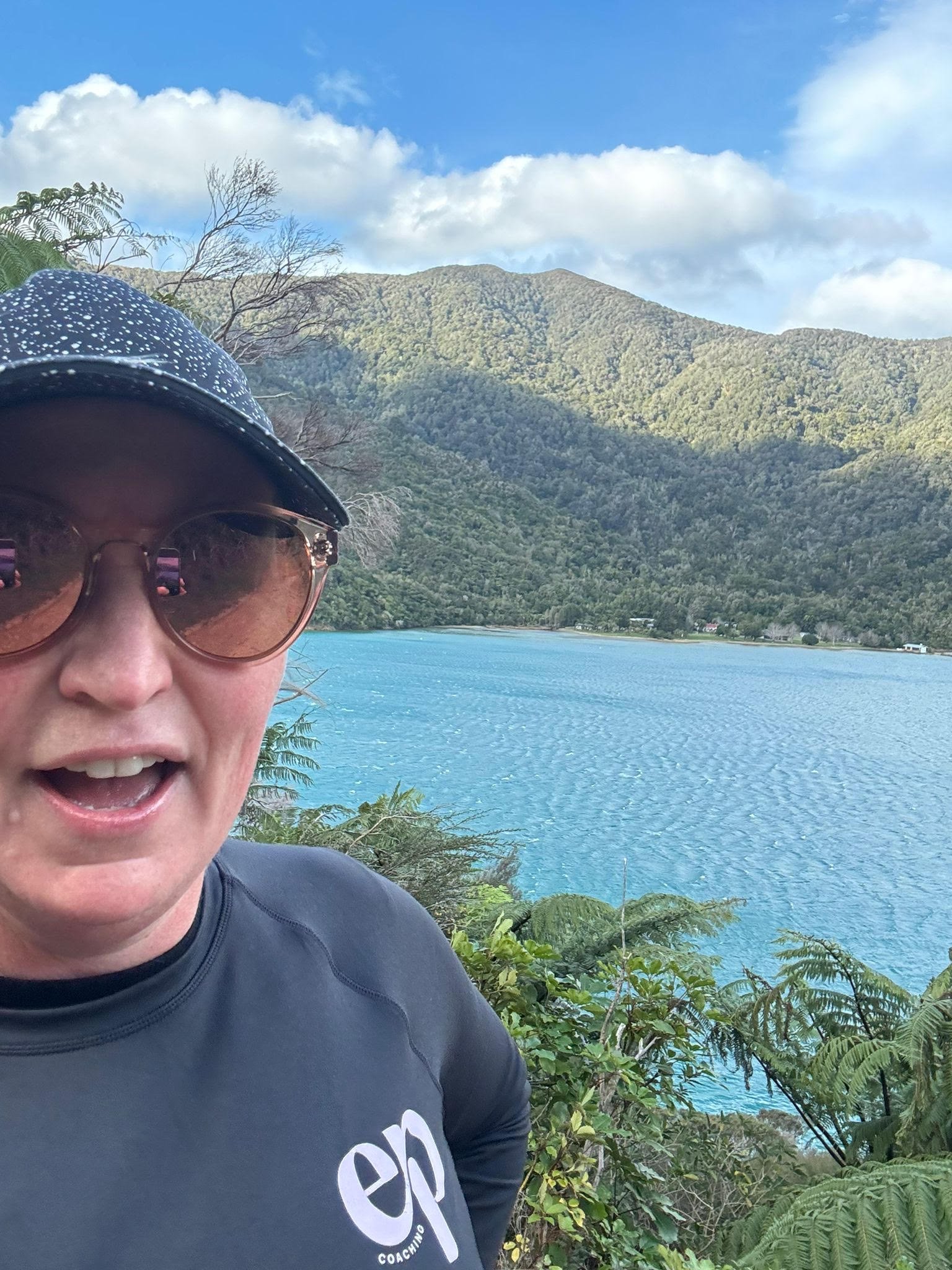 A woman wearing sunglasses and a cap taking a selfie with a scenic lake, green mountains, and a partly cloudy sky in the background.