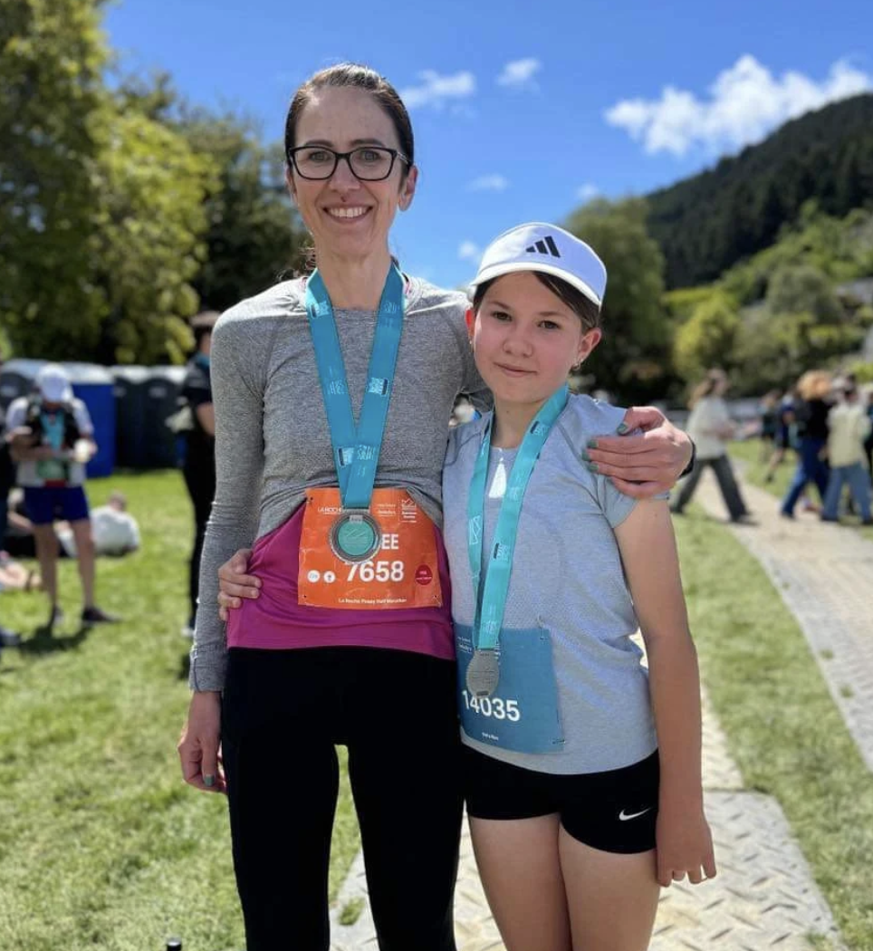 A woman and a young girl standing outdoors after a race, both wearing medals around their necks, with race bibs attached to their clothing. The woman has glasses and is smiling, while the girl is also smiling slightly. The background shows other people, trees, a blue sky with some clouds, and a scenic landscape.