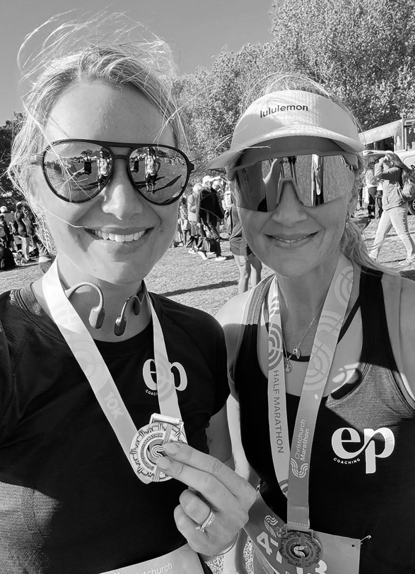 Two women smiling at the camera at a marathon event, both wearing sunglasses and medals around their necks, with a crowd and trees in the background.