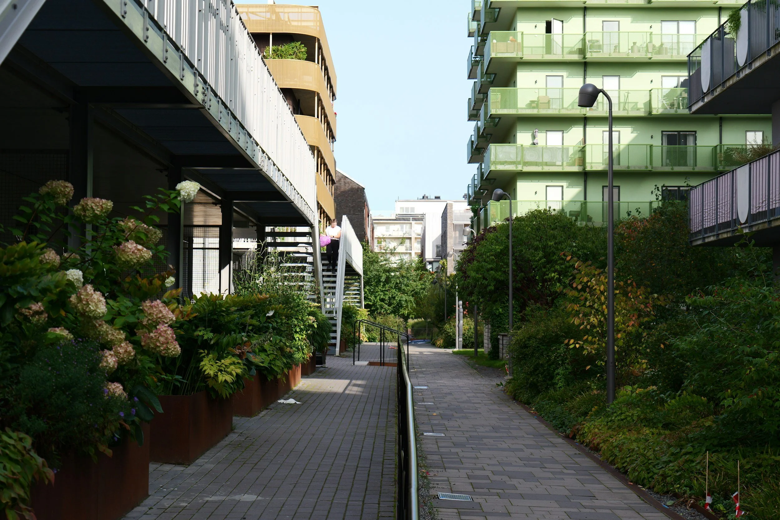 En travel gate i en boligbygning med grønne og beige balkonger, omgitt av blomster og grønne busker, under en klar blå himmel.