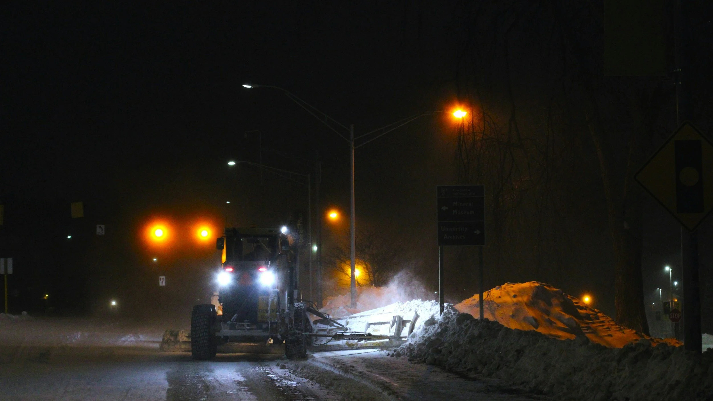 Snørydding av vei om natten med snøplog under dårlig utheving, lyser opp med blå og gule gate- og kjørelys og trafikklys.