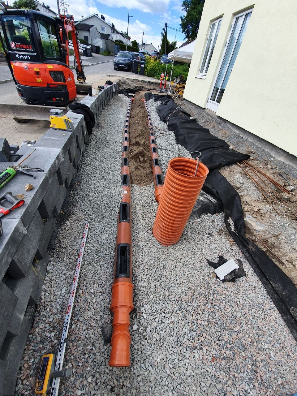 På en byggeplass er det gravd opp en vei med rørlegging. Det er en rød gravemaskin på venstre side, og avløpsrør ligger på bakken. Det er også svart geotekstilstoff og annet byggemateriale rundt gravingen.