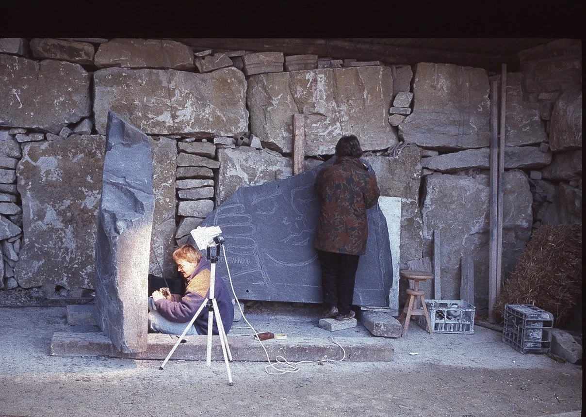 Two people working on stone carving outdoors, one sitting and one standing, with tools and equipment around them.