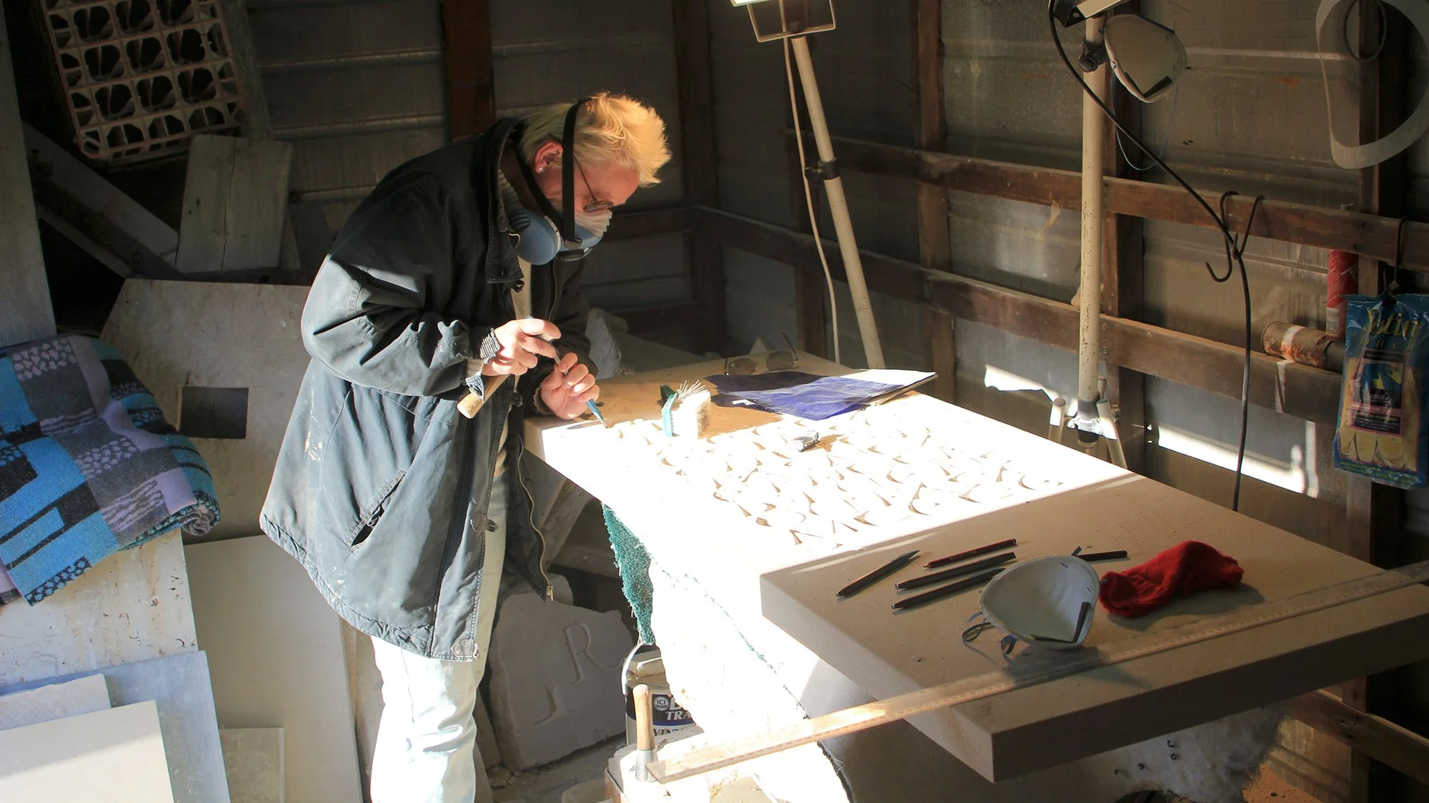Person working in a workshop, carving or shaping a piece of material on a large workspace lit by sunlight, surrounded by tools and materials.