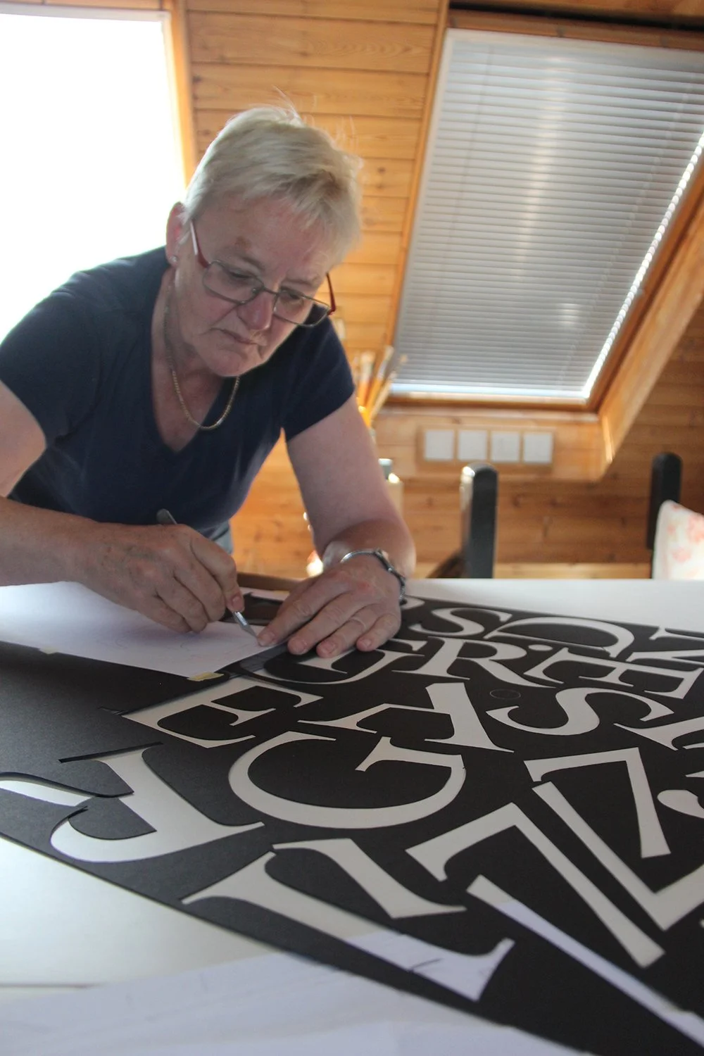An elderly woman with short blonde hair, glasses, and a black shirt, is working on black and white cut-out letters at a table inside a wooden room with sloped ceiling and window blinds.