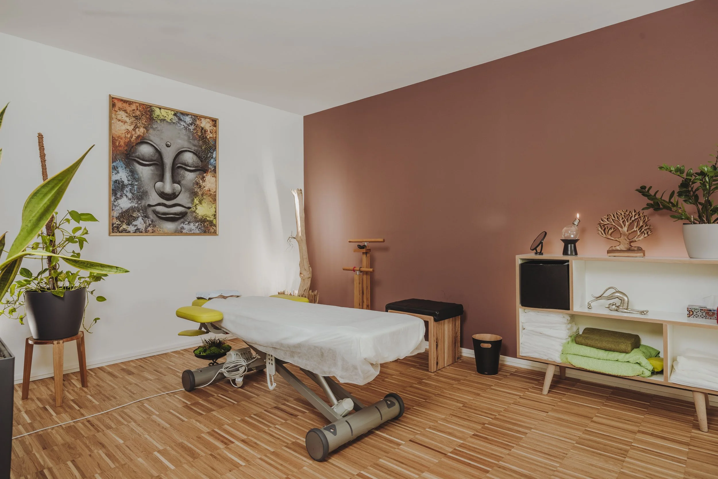Ein Behandlungsraum mit Massagetisch, Pflanzen, Wandkunst eines Buddha-Gesichts, und Möbeln in neutralen Farben.