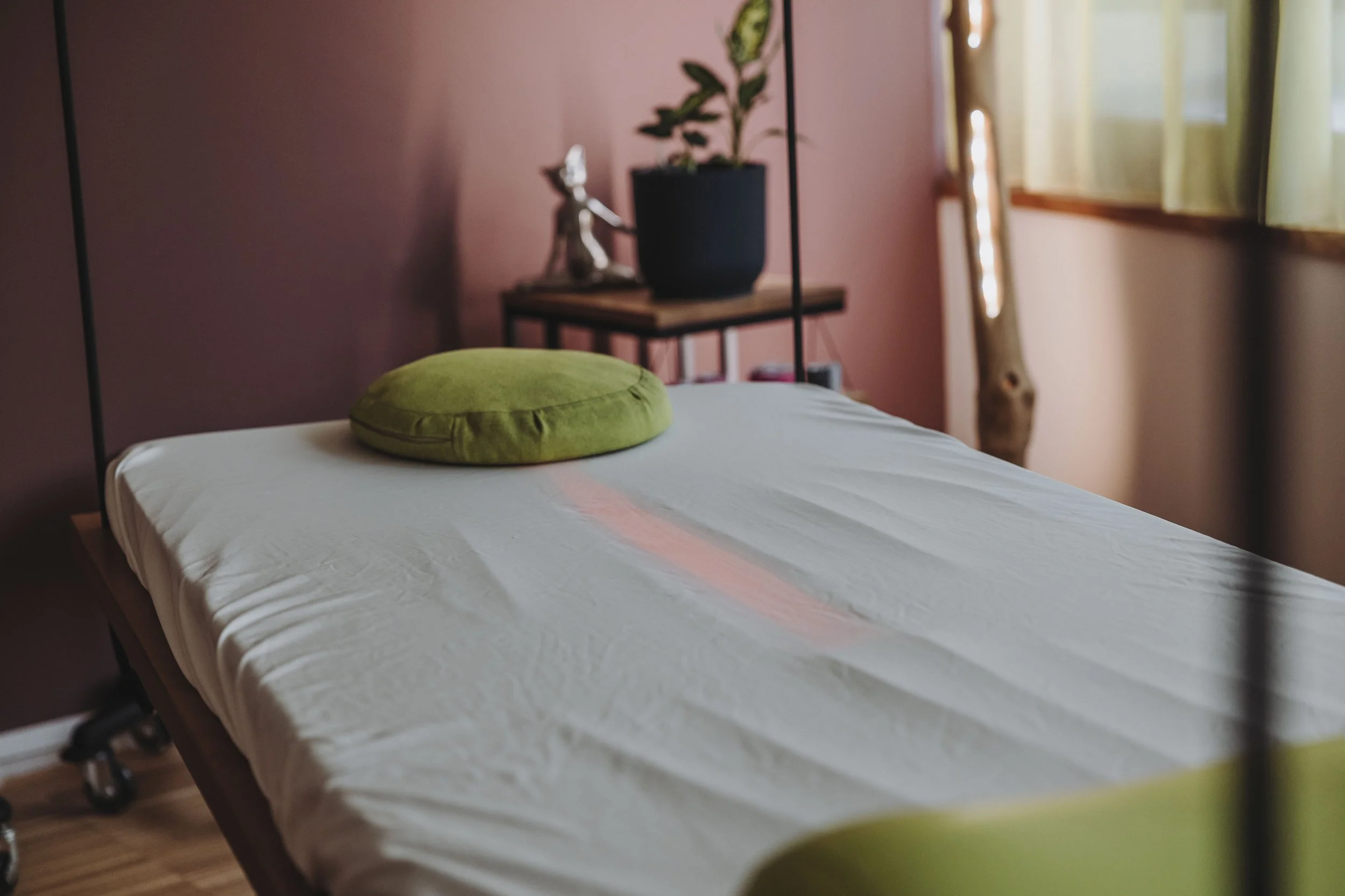 Eine Therapieliege mit einem grünen Kissen im Behandlungsraum. Im Hintergrund ein kleiner Tisch mit Pflanze und Dekoration, neben einem Fenster mit grünen Vorhängen.