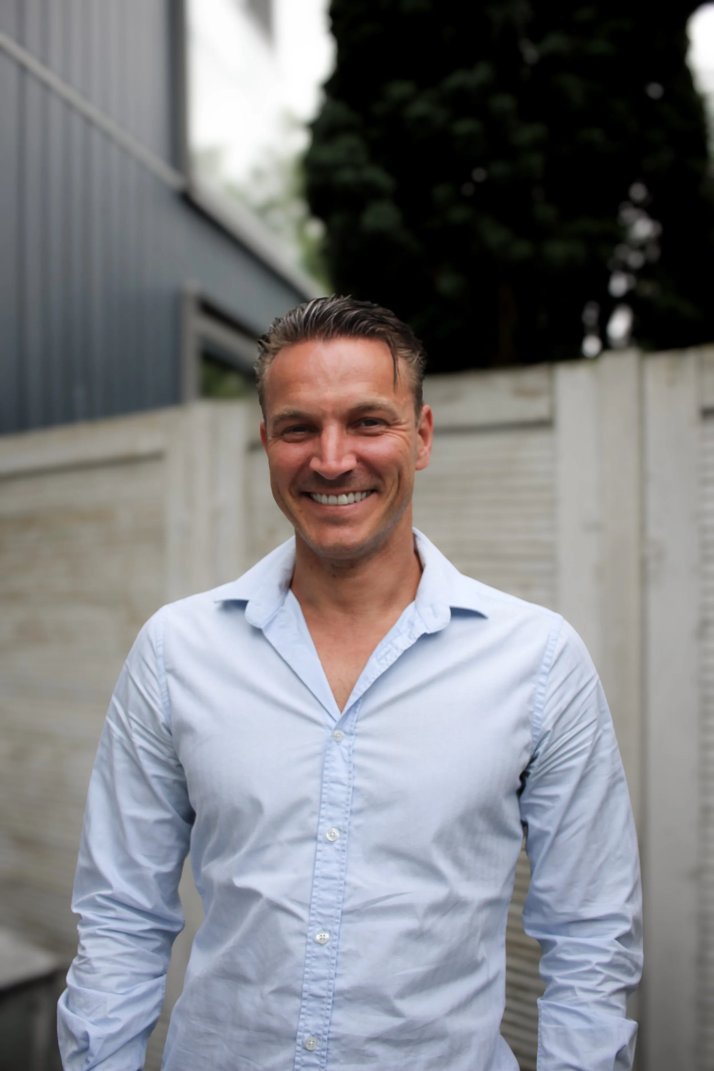 Man (Michiel Muller) glimlachend in een tuin met een witte schutting en bomen op de achtergrond.