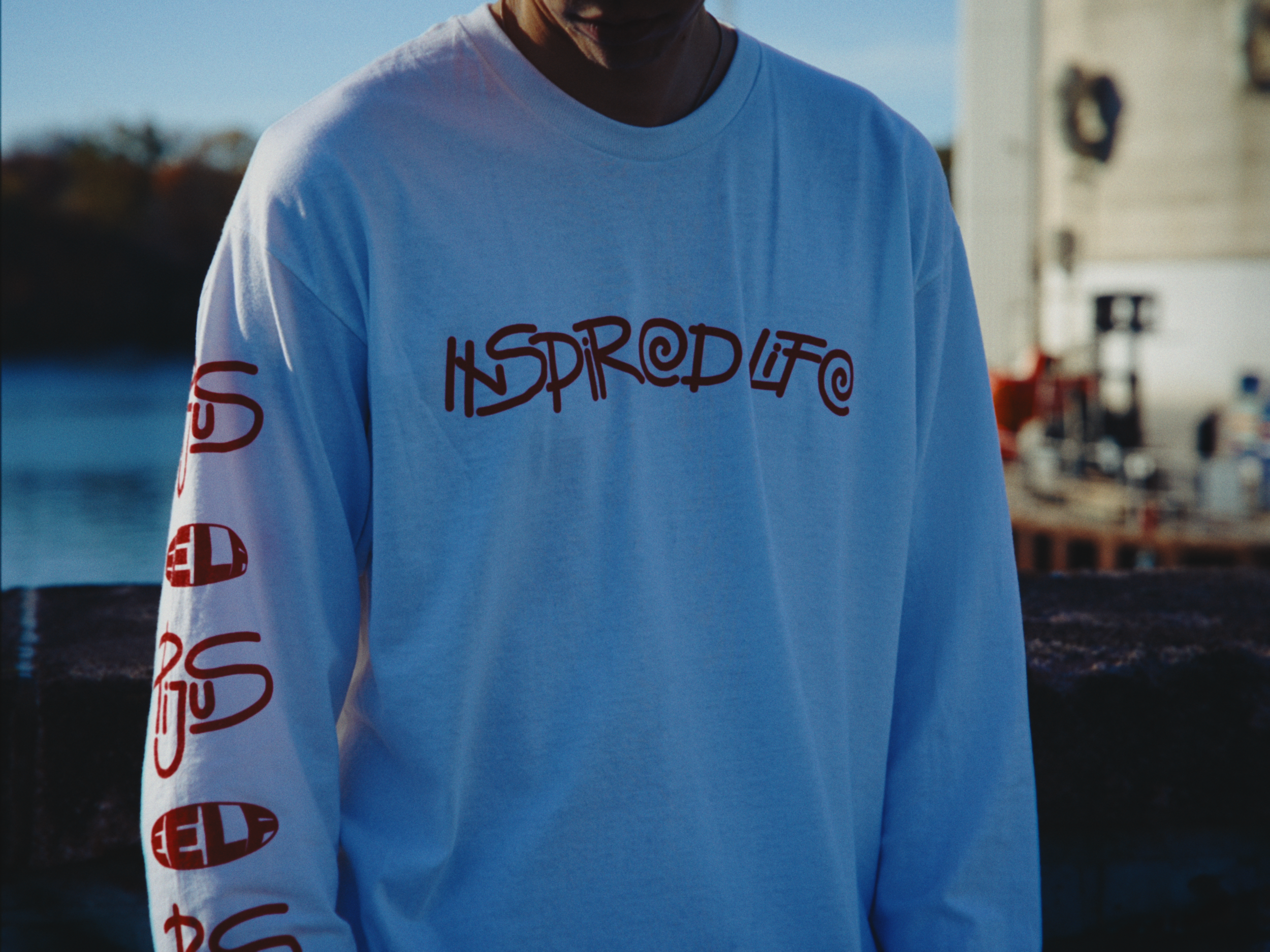 Person wearing a light blue long-sleeve shirt with the words 'Inspired Life' written on the front and the word 'GEL' repeated on the sleeve, standing outdoors near water, with buildings in the background.