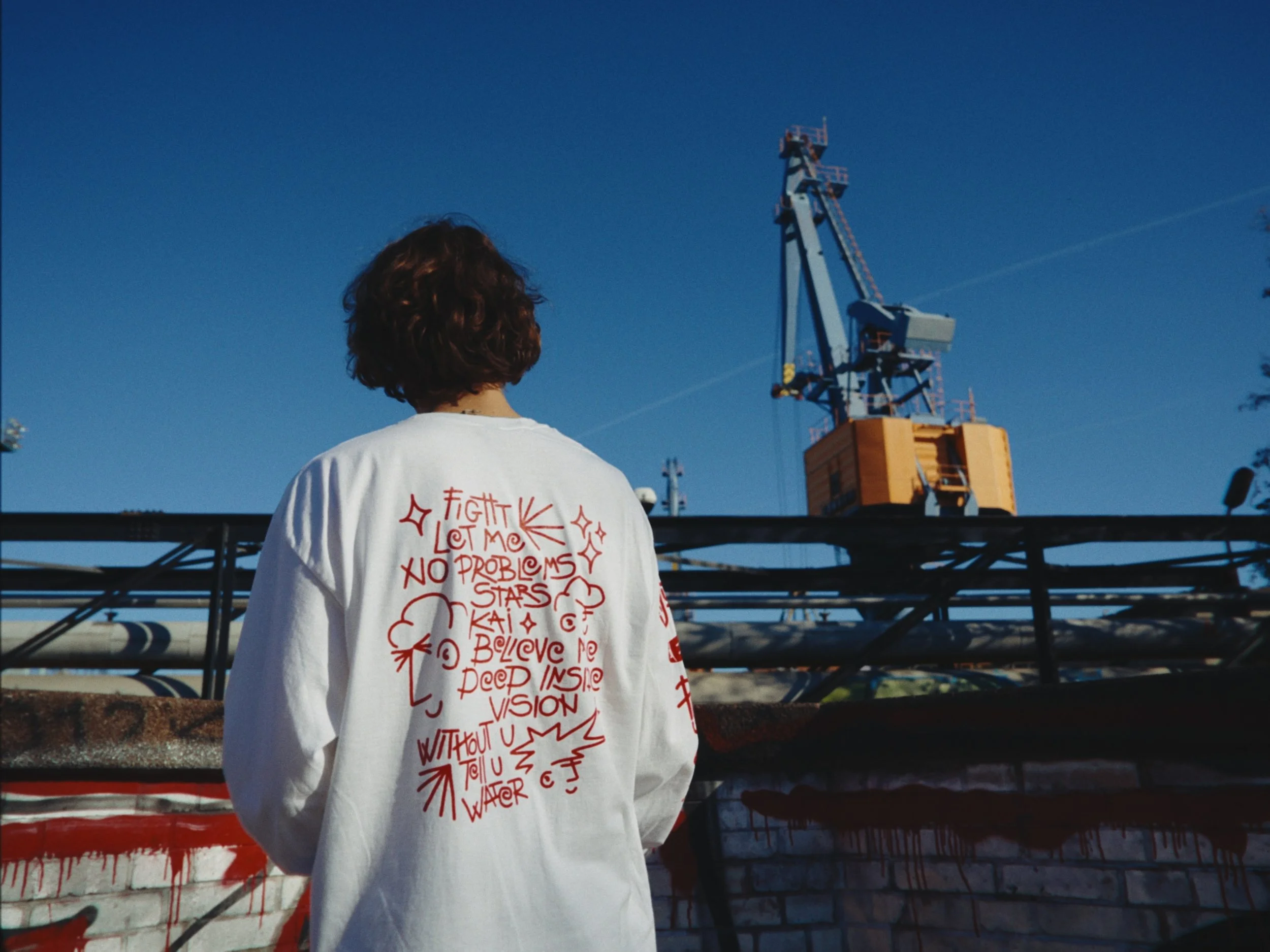 Person with curly hair wearing a white shirt with red writing and drawings, facing away from the camera, standing outdoors with industrial equipment and a blue sky in the background.
