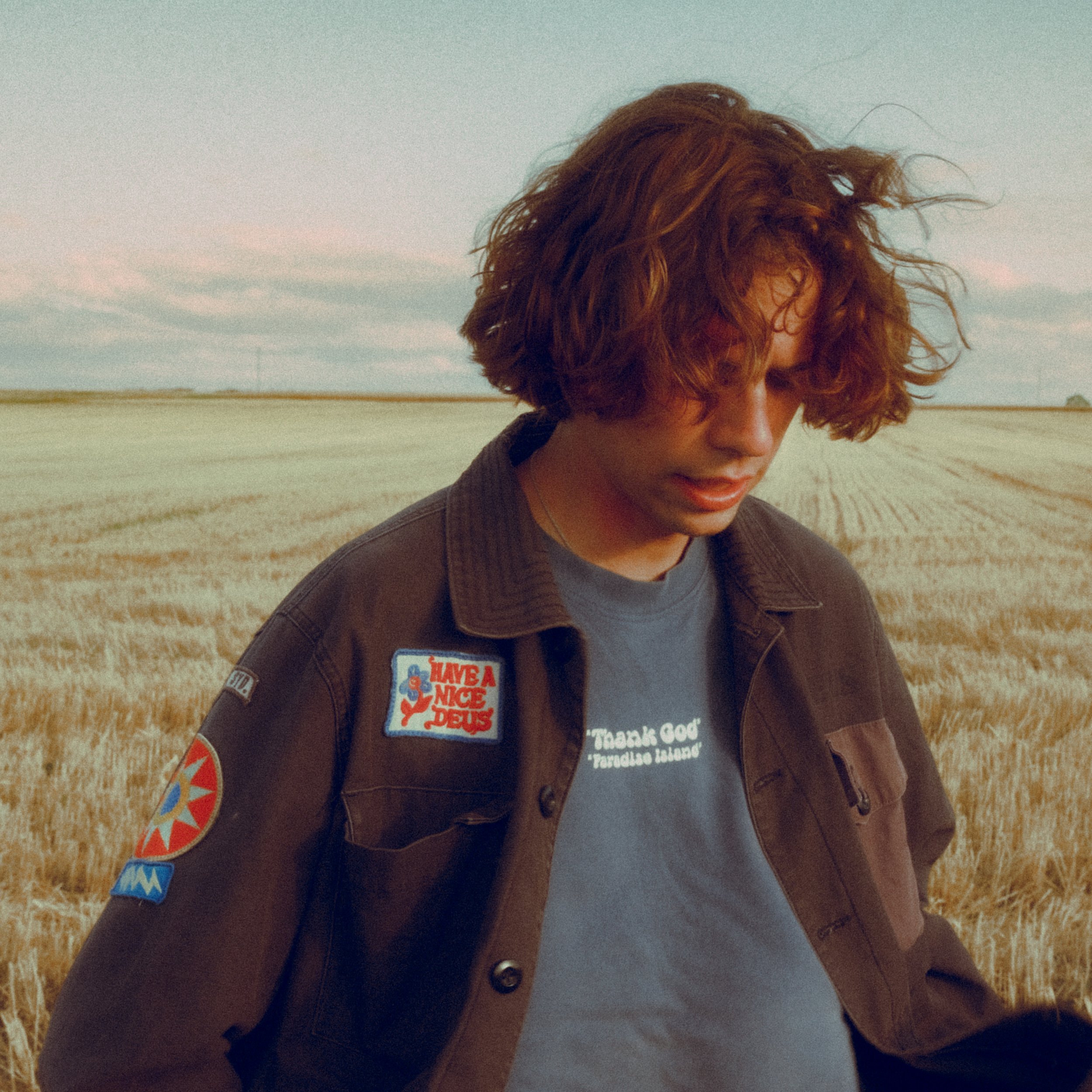 A young man with curly red hair looking down in a wheat field during daytime, wearing a brown jacket with patches and a gray T-shirt.