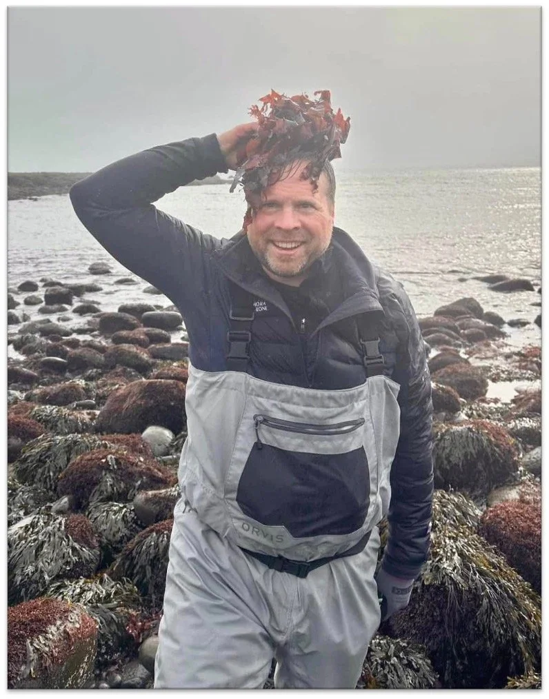 A smiling man in waterproof outerwear standing on a rocky shoreline holding a bunch of wet red leaves on his head.