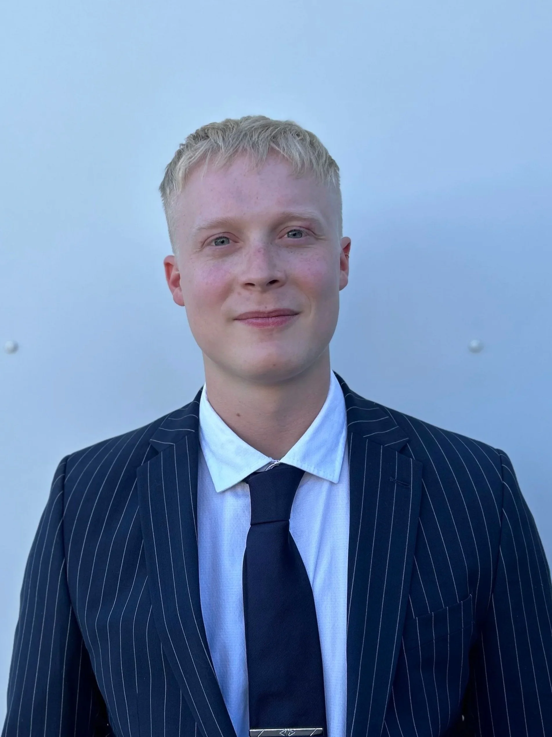 A young man with blonde hair, wearing a dark pinstripe suit, white shirt, and dark tie, standing against a light blue background.