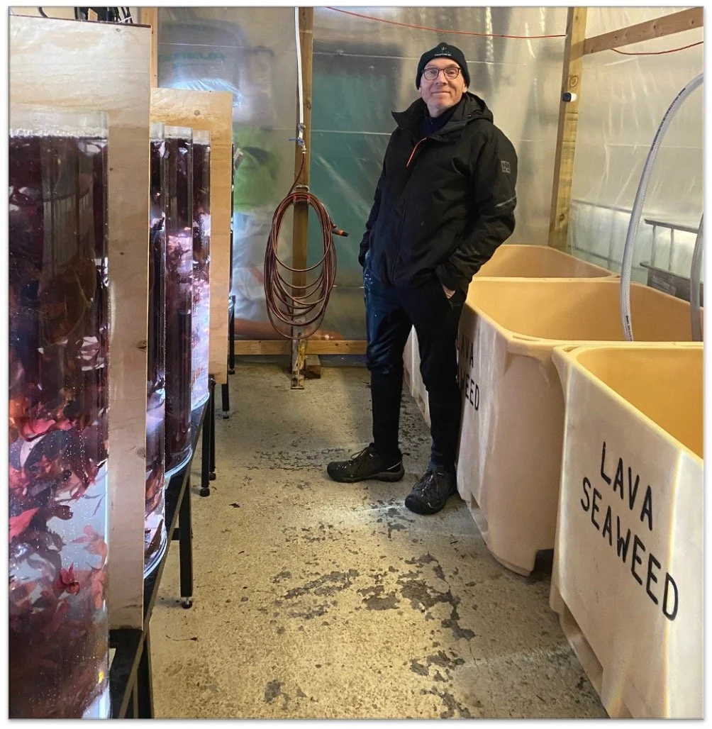 A man wearing glasses and a black jacket stands inside a seafood farm or shellfish hatchery, surrounded by tanks labeled 'LAVA SEAWEED' and 'OYSTERS.' He is smiling and hands in pockets, with industrial equipment and hoses visible in the background.