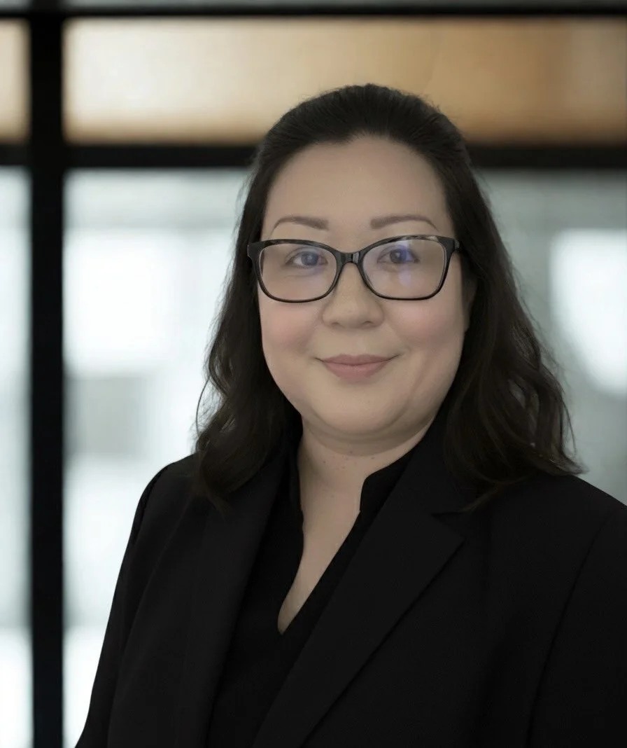 A woman with shoulder-length black hair, wearing glasses, a black blazer, and a black shirt, standing indoors with a blurred window or glass wall in the background.