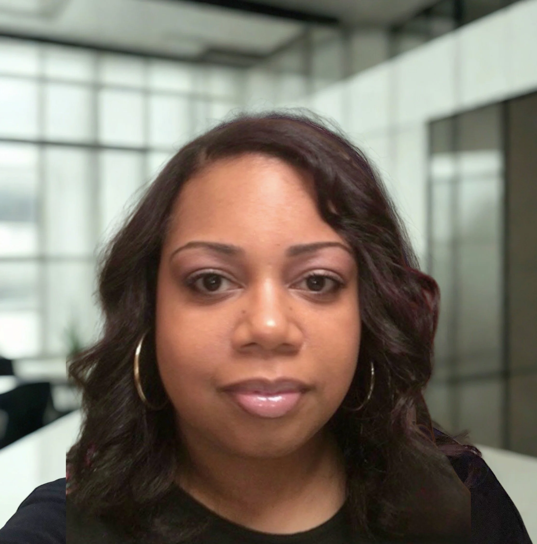 Close-up photo of a woman with shoulder-length dark hair, wearing hoop earrings and a black top, in a modern office space with large windows.
