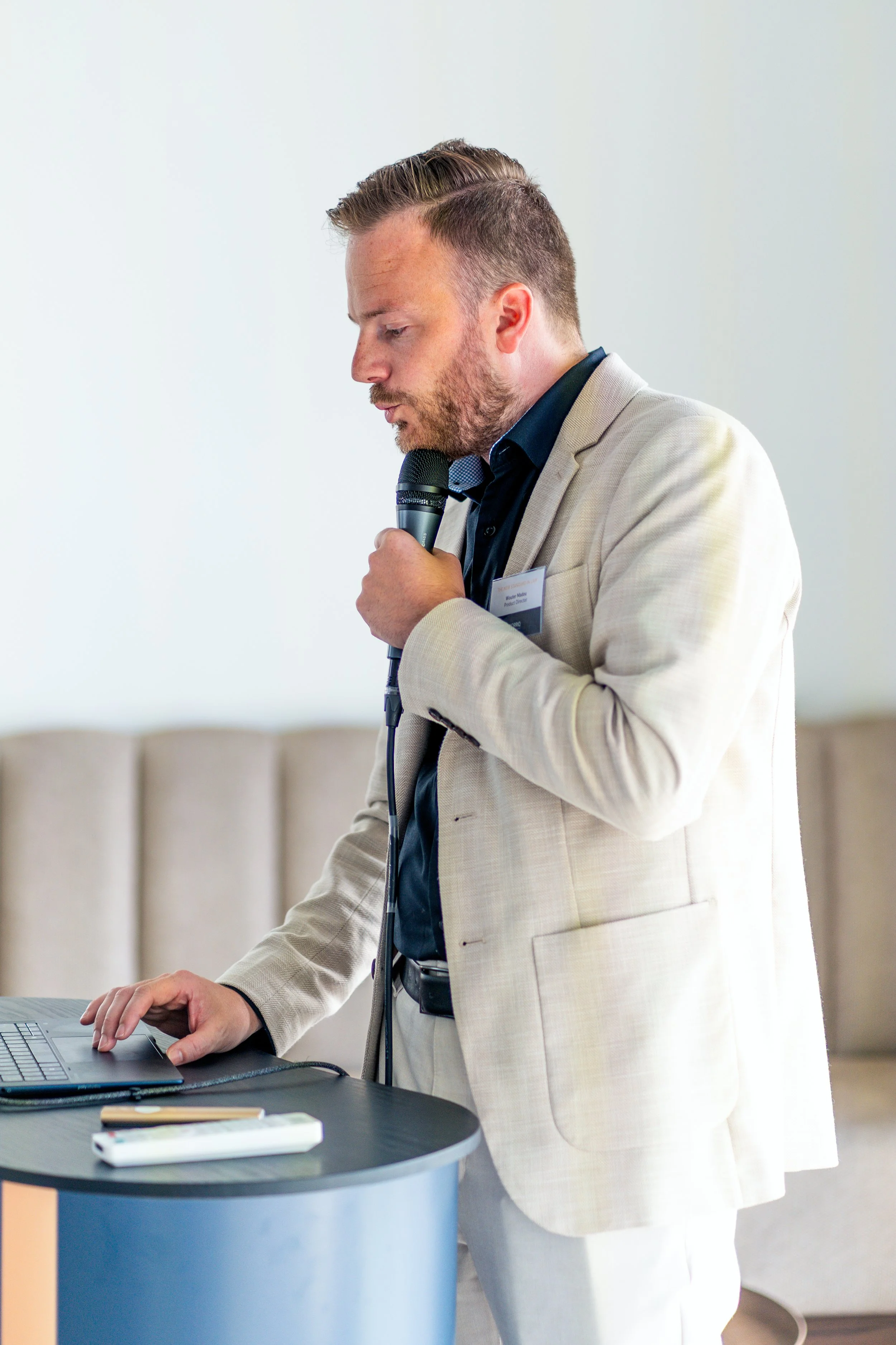 A man in a beige blazer and white pants, holding a microphone, appears to be speaking or presenting at a formal event. He is standing next to a table with a laptop and some office supplies, in a bright room.