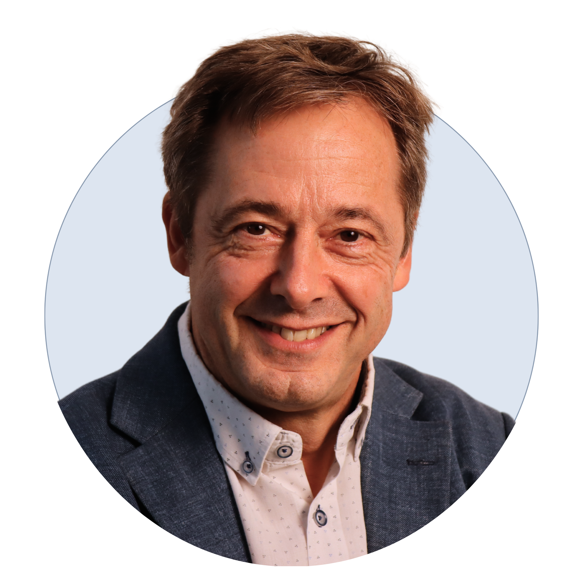 Close-up of a smiling man with light skin, brown hair, wearing a dark blazer and a white shirt with small dark patterns.