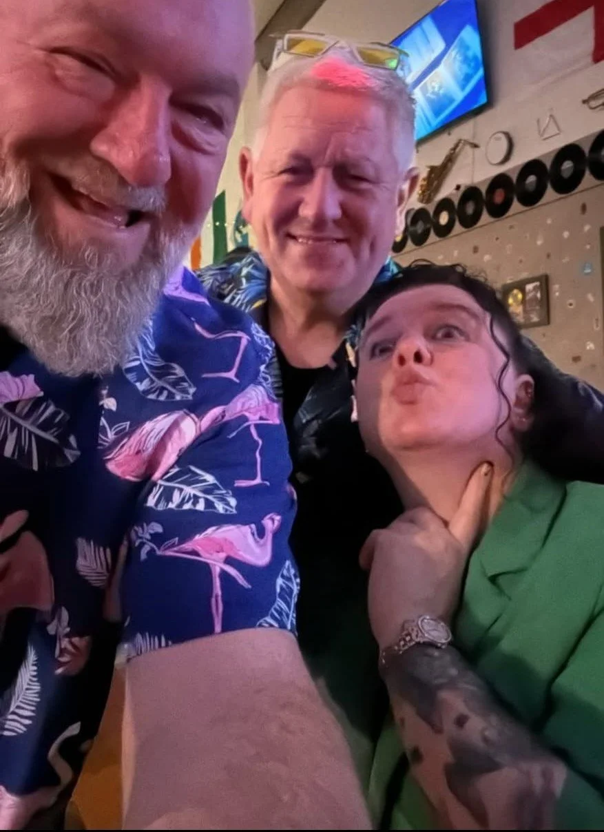 Three people posing for a selfie in a social setting, smiling, with a bar or restaurant background, and a television screen on the wall.