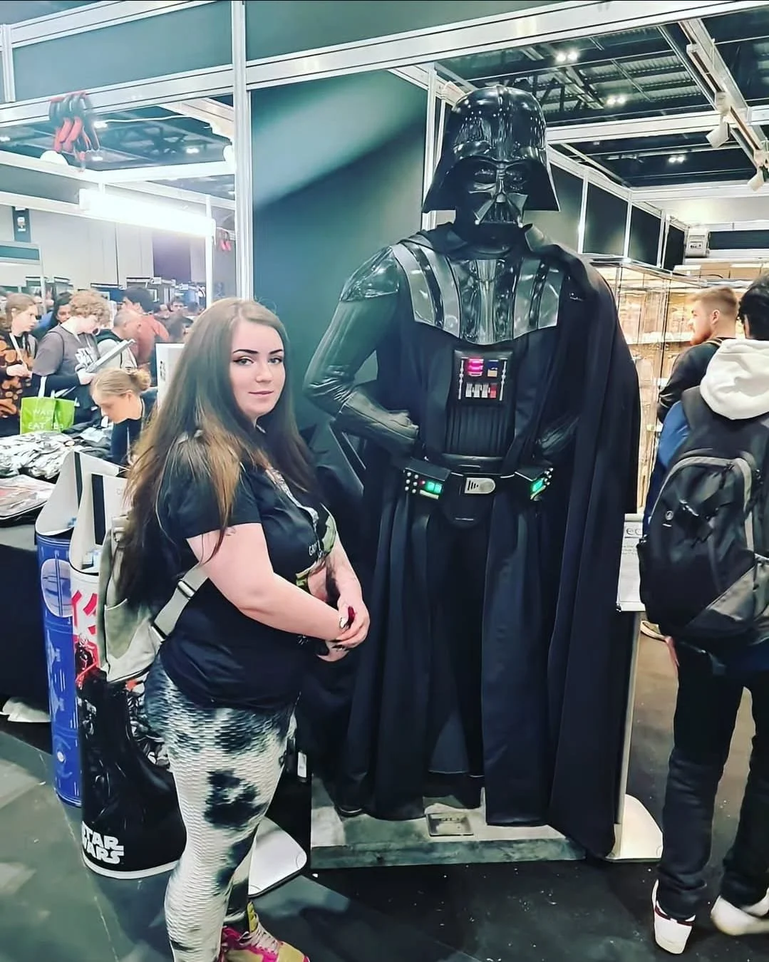 A young woman with long brown hair standing next to a life-sized Darth Vader costume at a convention or expo.