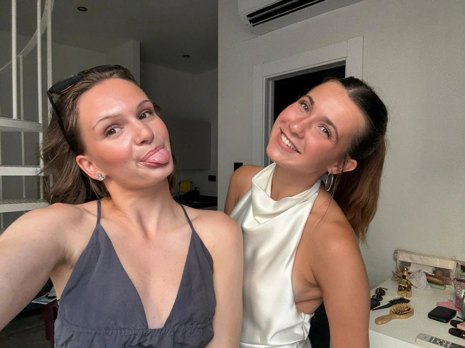 Two women taking a selfie indoors, one making a funny face with her tongue out, the other smiling brightly. The woman on the left has light brown hair, sunglasses on her head, and is wearing a dark gray spaghetti strap top. The woman on the right has dark brown hair, hoop earrings, and is wearing a white satin sleeveless top.