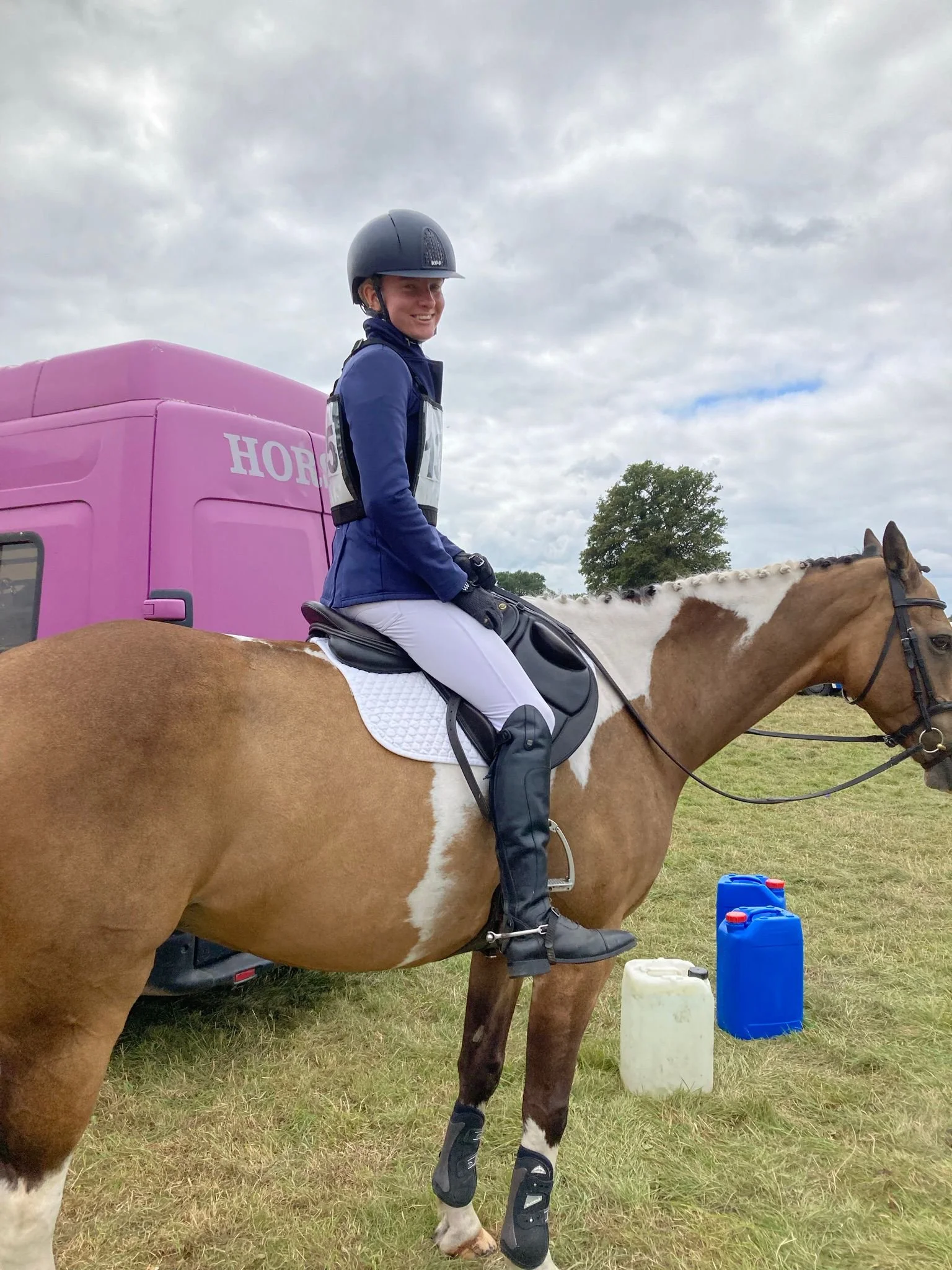 A woman wearing a helmet and riding gear sits on a brown and white pinto horse in a grassy field, with a pink trailer and water containers nearby, under a cloudy sky.