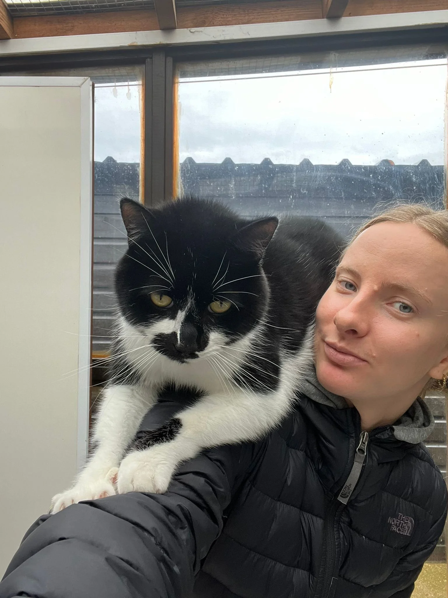 A woman in a black North Face jacket taking a selfie indoors with a black and white cat perched on her shoulder, against a background of windows and a cloudy sky.