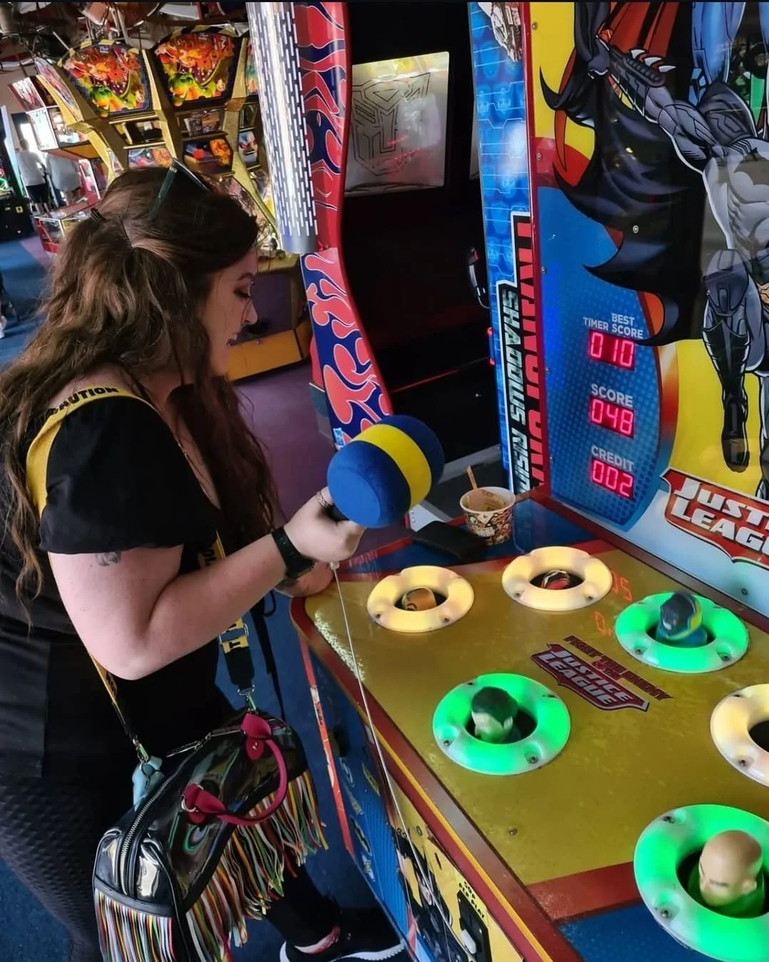 Young woman playing a carnival game where she uses a hammer with a soft mallet to hit a metal plate with a ring on it. The game is themed after 'The League of Legends' and has a colorful, superhero comic-style design.