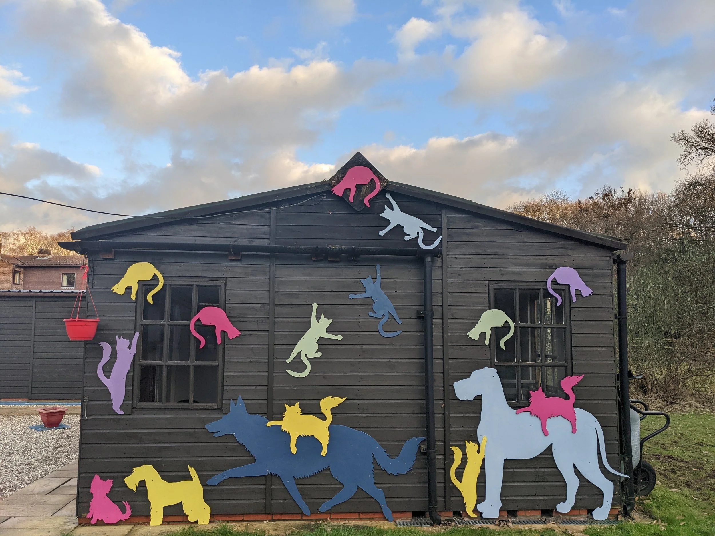 Colorful animal cutouts, including cats, and dogs, decorate a dark wooden house exterior against a cloudy sky.
