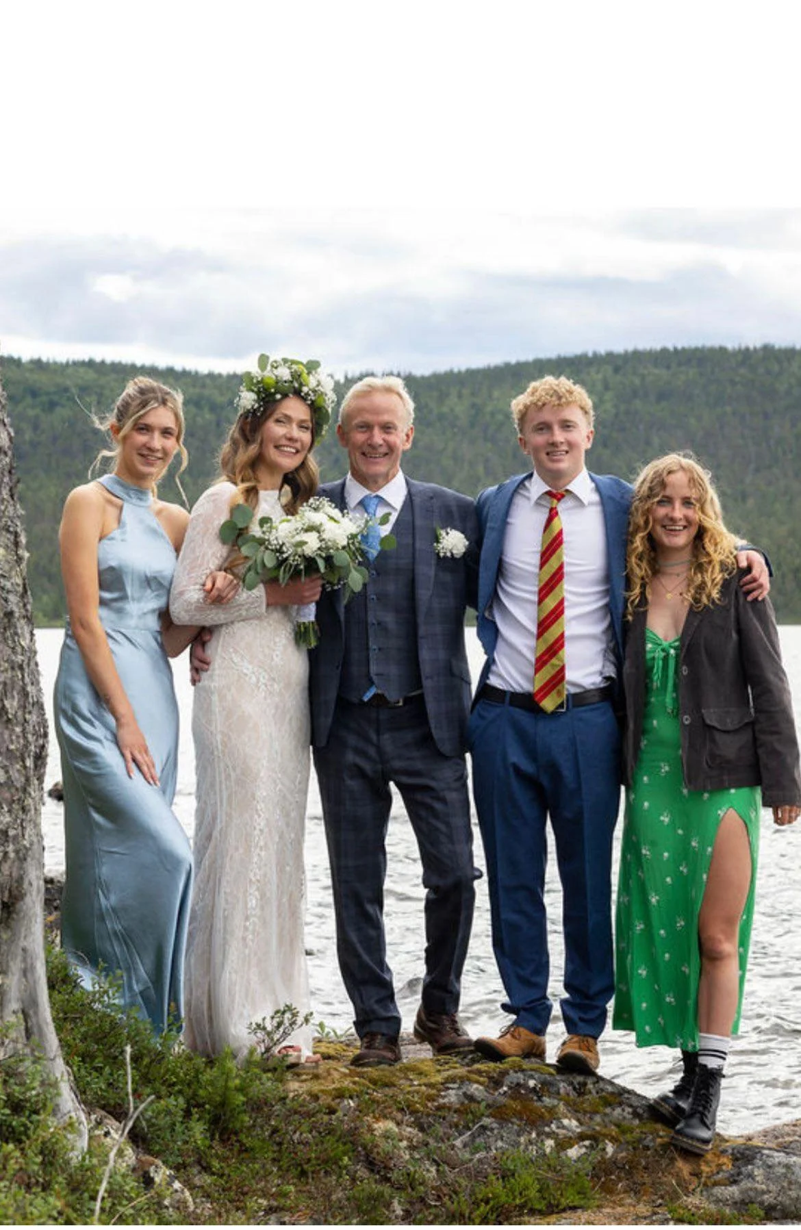A family of five celebrating outdoors near a lake with a green forested mountain background. The woman in the center is wearing a white lace wedding dress and a flower crown, holding a bouquet. The man next to her wears a checkered suit with a boutonniere. The others are dressed in colorful semi-formal attire, smiling and posing together.