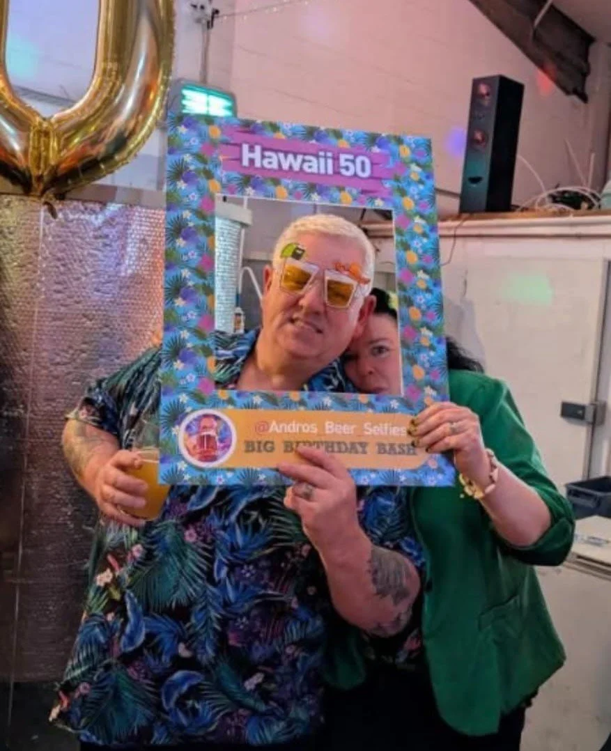 A man with blonde hair and sunglasses is smiling and holding a drink, standing behind a colorful party frame that says 'Hawaii 50'. A woman in a green jacket is next to him, partially visible, holding the frame. The background indicates a party or celebration setting.