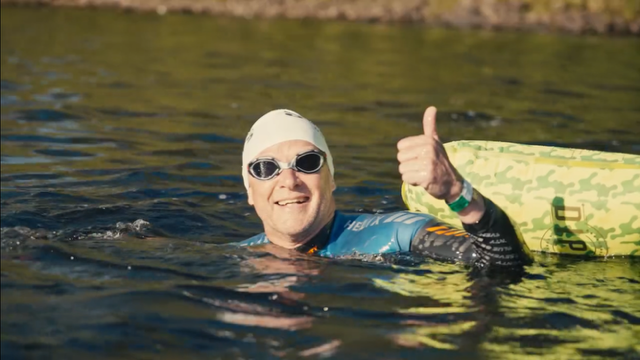 An older woman swimming in a body of water, wearing goggles, a swim cap, and a wetsuit, giving a thumbs-up sign and smiling.