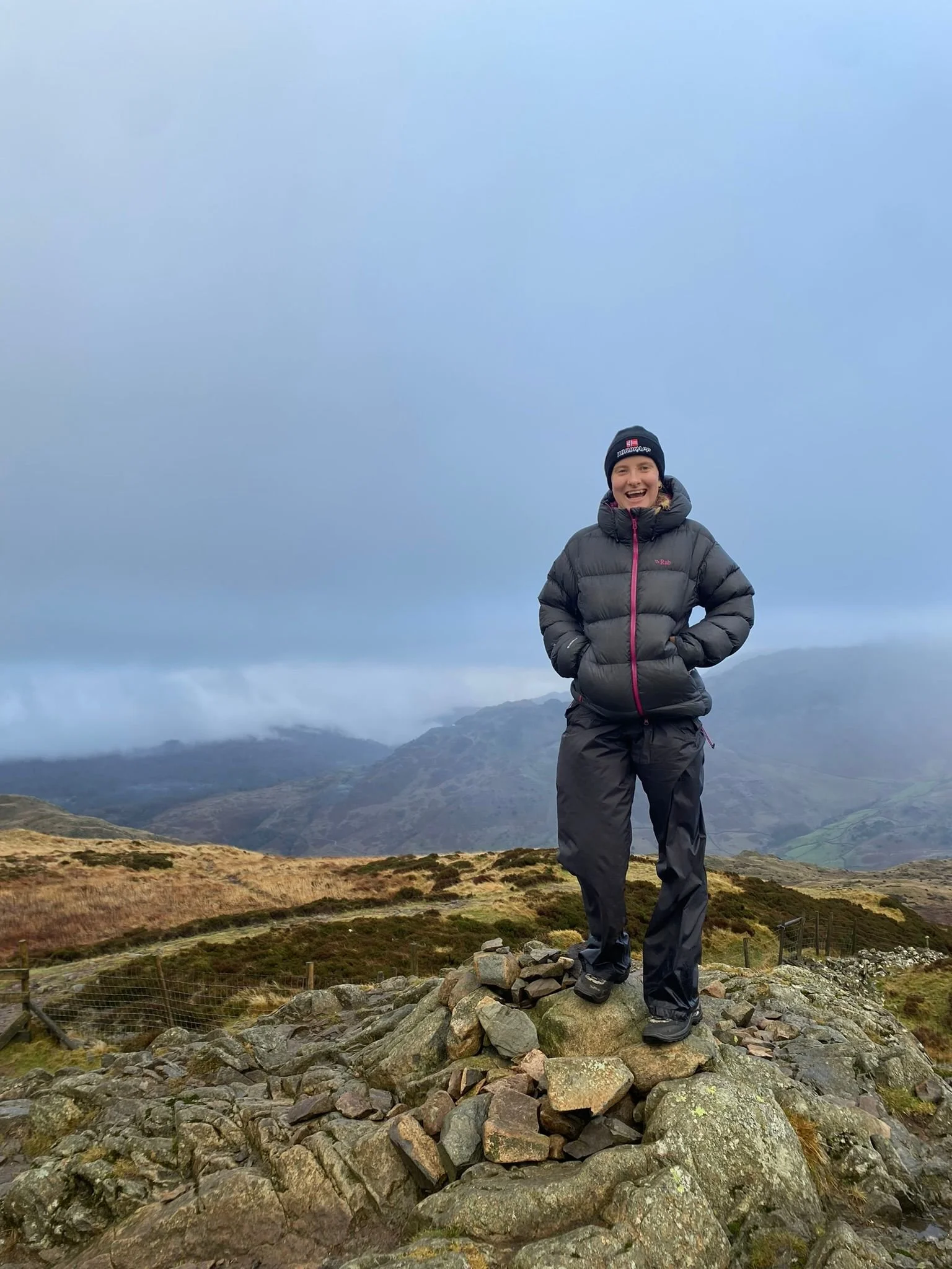A person standing on a rocky hilltop in a mountainous landscape, wearing a black beanie, a gray puffy jacket, and black pants, smiling at the camera with mountains and a cloudy sky in the background.
