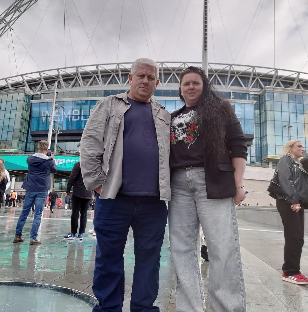 Two people standing outside Wembley Stadium in London, with the stadium in the background. It is cloudy and wet.