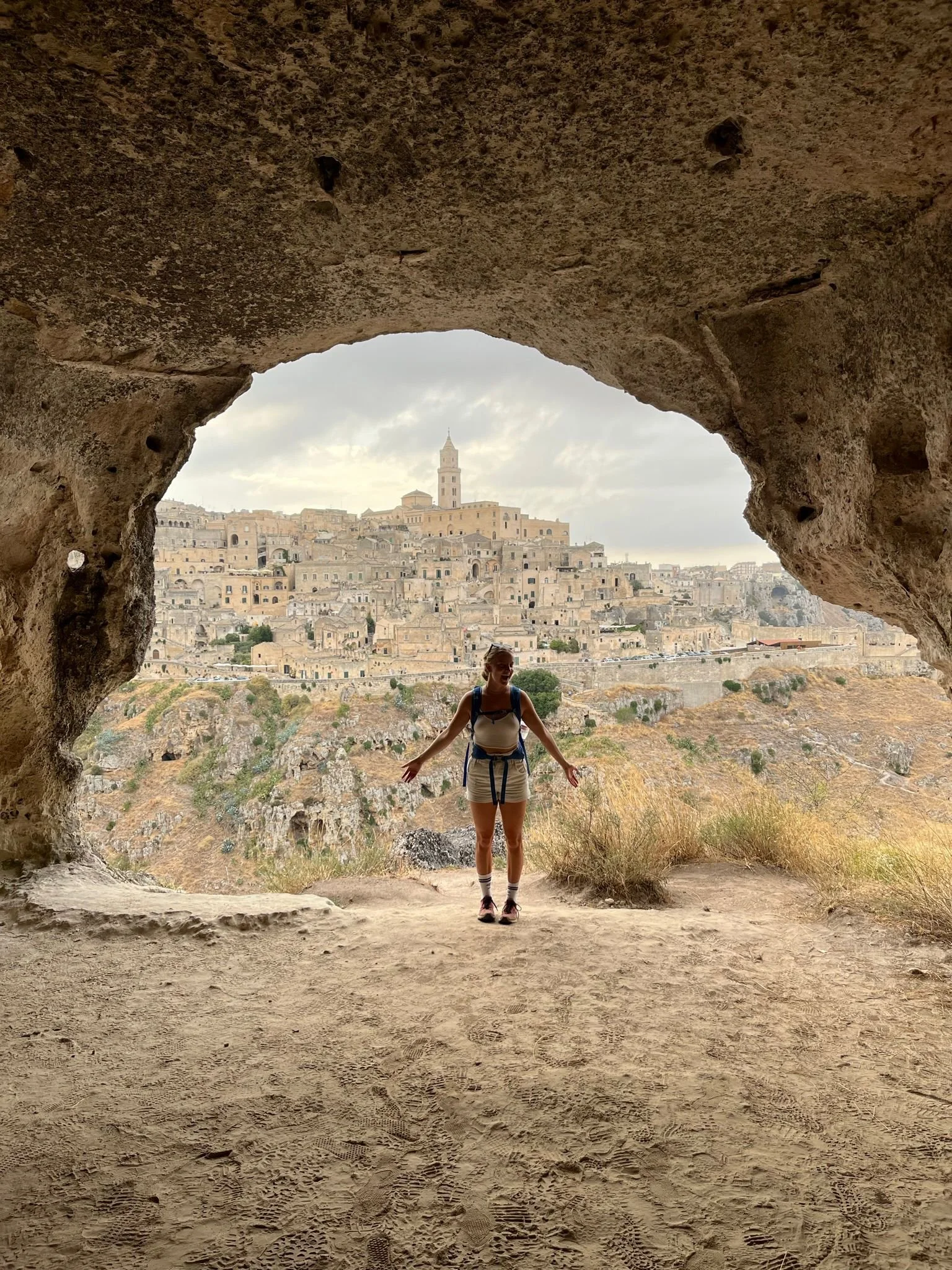 A woman standing in front of a cave opening, with an ancient hillside town and a tall tower in the background.