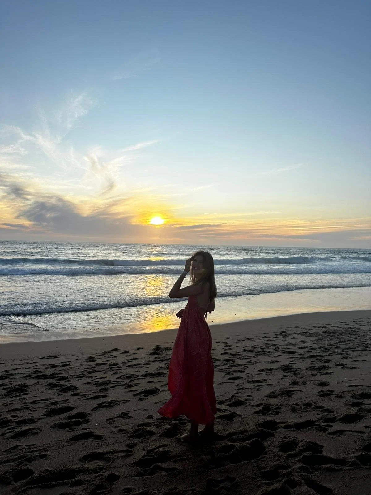 A woman in a red dress standing on a sandy beach at sunset, with the ocean and sky in the background.
