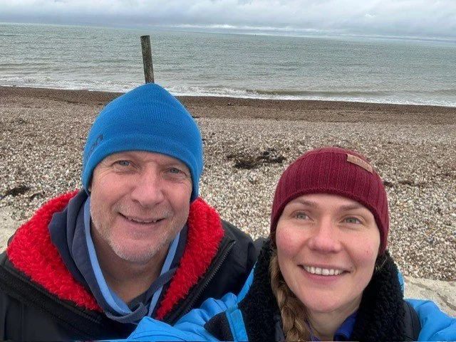 A man and woman smiling at the camera on a pebbled beach with the ocean and cloudy sky in the background. They are dressed in warm clothing, including beanies.