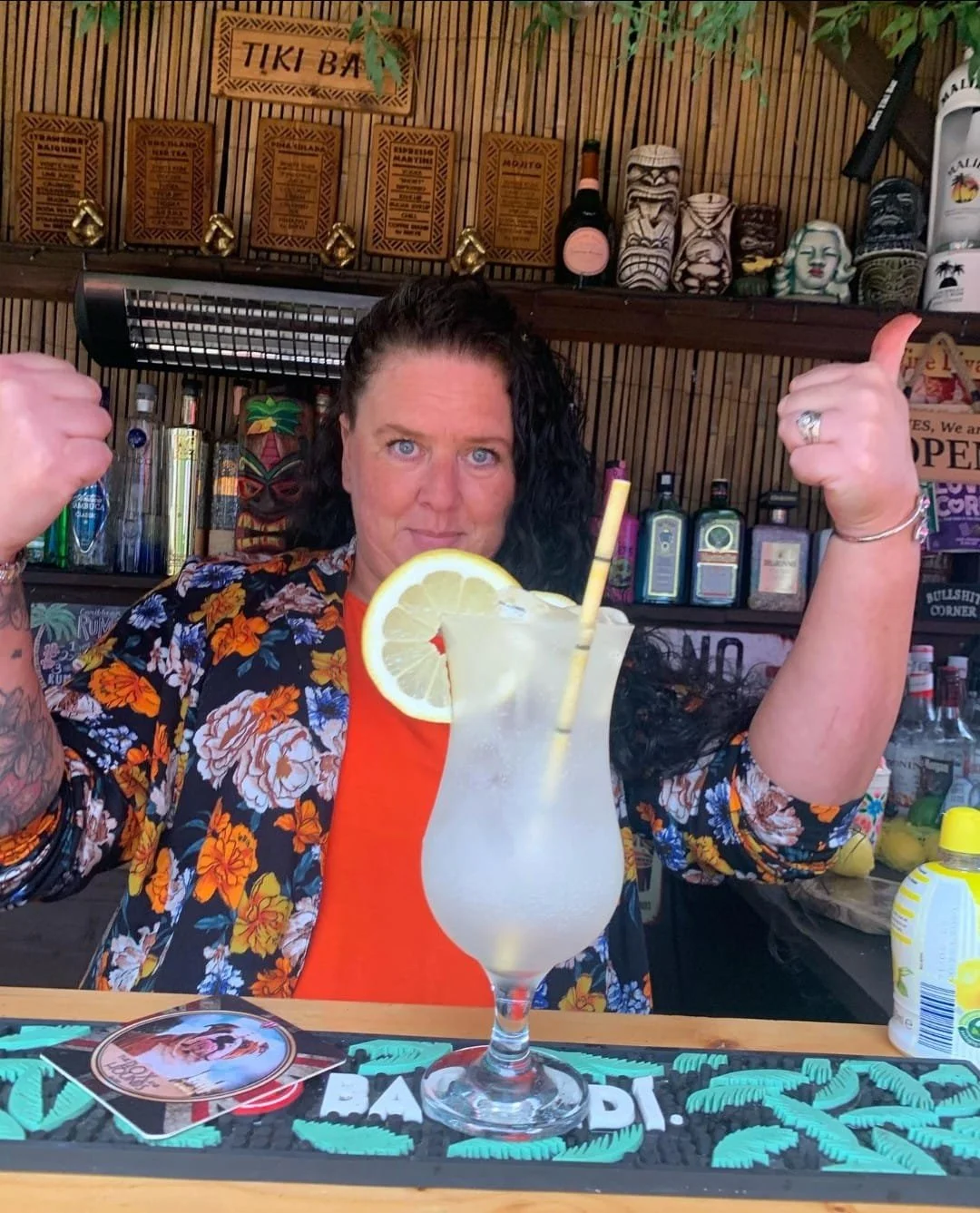 Woman with curly black hair and blue eyes at a tiki bar, wearing a floral shirt and orange top, giving two thumbs up. In front of her is a large tropical cocktail with lemon slice and straw. The background has tiki masks, bottles, and a wooden sign that says 'TIKI BAR'.