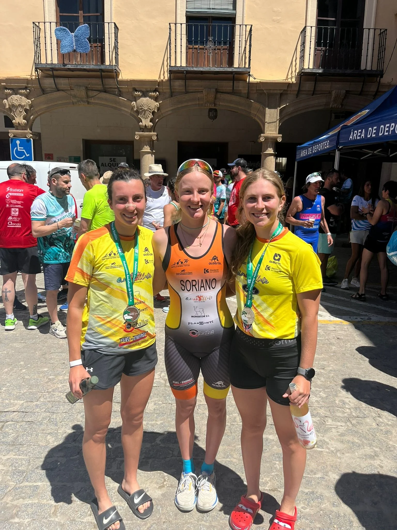 Three young women standing together outdoors after a race, wearing athletic gear and medals, with a crowd in the background.