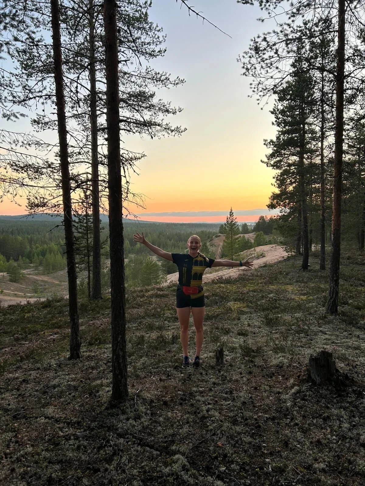 A person standing in a forest clearing with arms outstretched, smiling, during sunset, with trees and a distant horizon in the background.