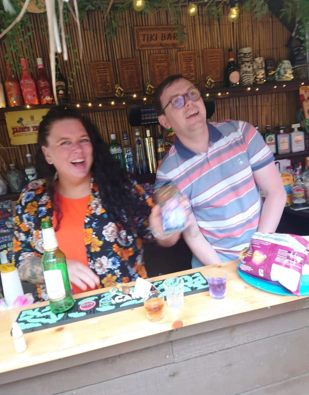 Two people behind a bar counter at a tiki bar, smiling and holding drinks, with bottles and tiki masks in the background.