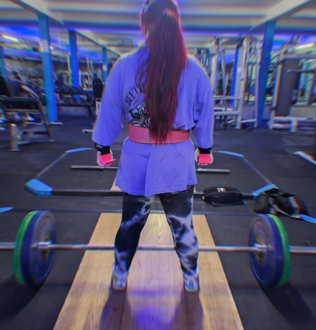A woman with long hair in a gym, standing in front of a barbell on a wooden platform, preparing to lift weights.